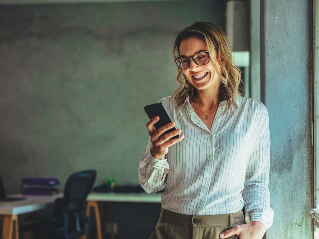 Nainen seisoo toimistoympäristössä ja hymyilee katsoessaan älypuhelintaan, pukeutuneena vaaleaan puseroon ja ruskeisiin housuihin.